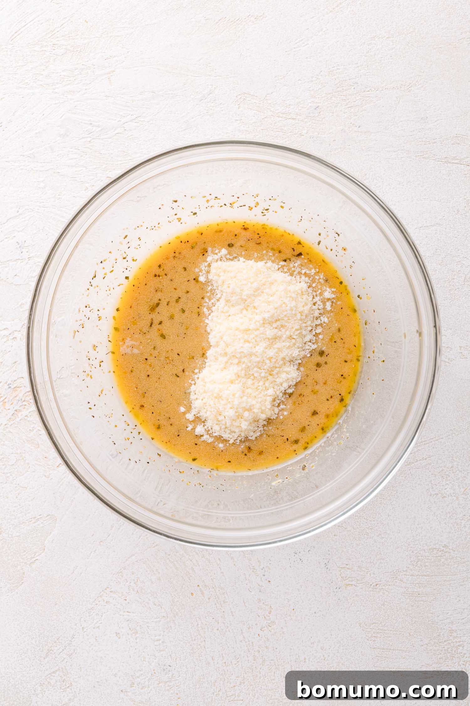 A clear glass bowl containing a fully mixed dressing, topped with a generous mound of finely grated Parmesan cheese, ready to be stirred in, all set on a light-colored surface.