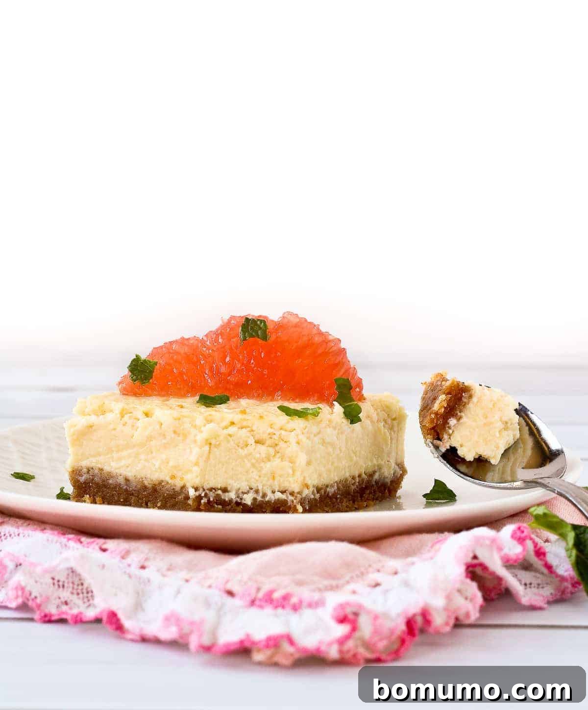 A close-up of a grapefruit cheesecake square with a bite removed, revealing the creamy texture and crumbly crust, garnished with fresh mint.