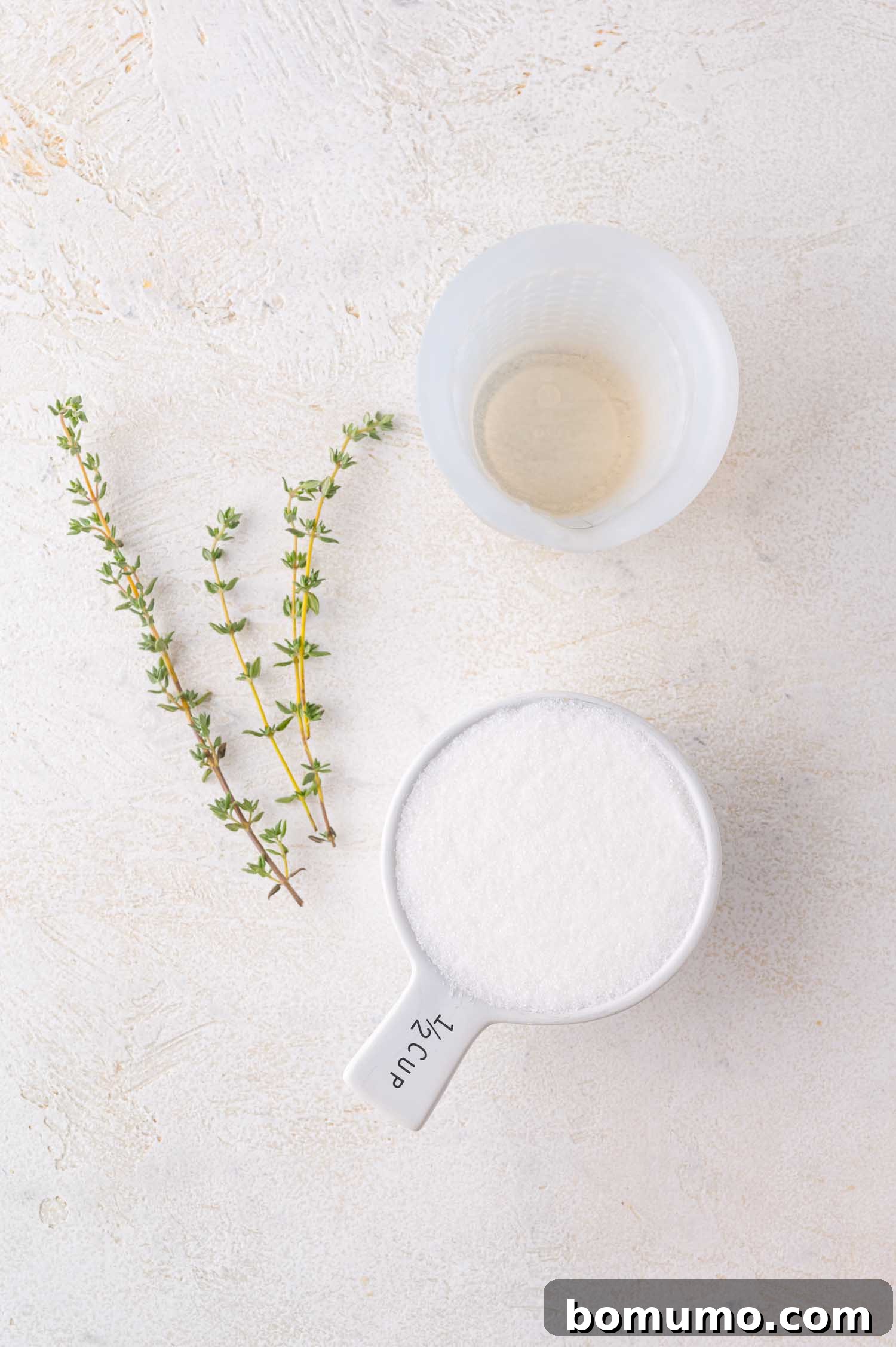 A visual of half a cup of granulated sugar, a small clear cup of water, and two sprigs of fresh thyme, neatly arranged on a light-colored surface, demonstrating the initial ingredients for the simple syrup.