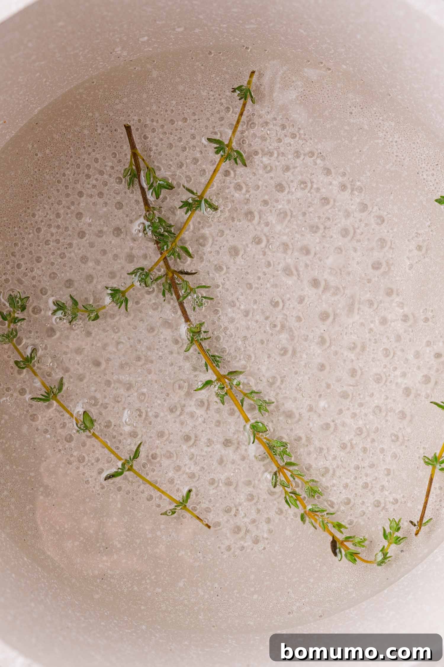 Close-up of two fresh thyme sprigs gently floating in a saucepan filled with simmering simple syrup, illustrating the infusion process.