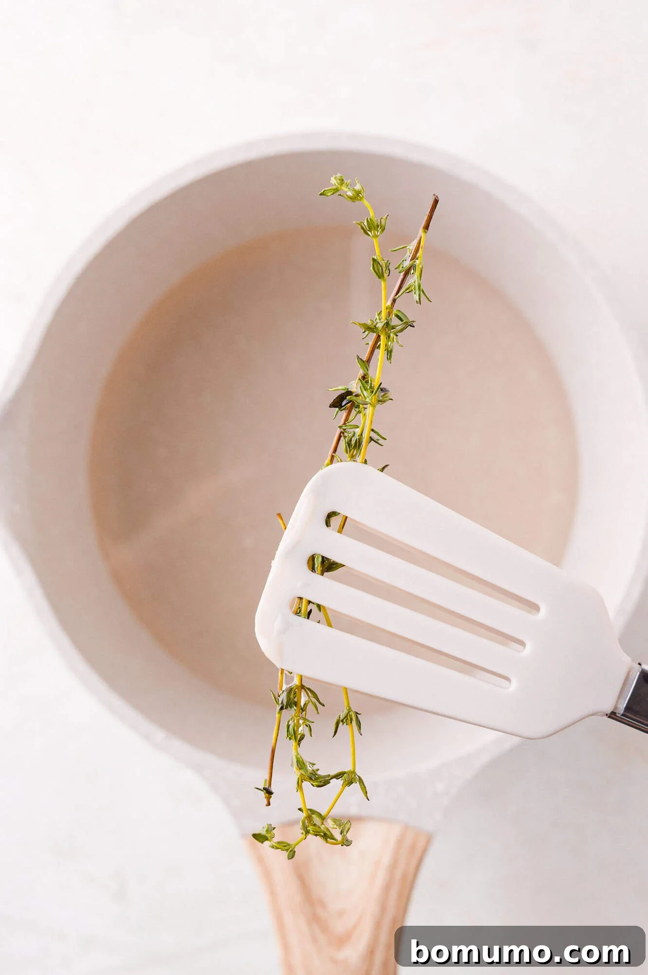 A white spatula holds a steeped sprig of fresh thyme over a white saucepan, containing the now infused simple syrup. This image captures the moment before the thyme is removed from the syrup.