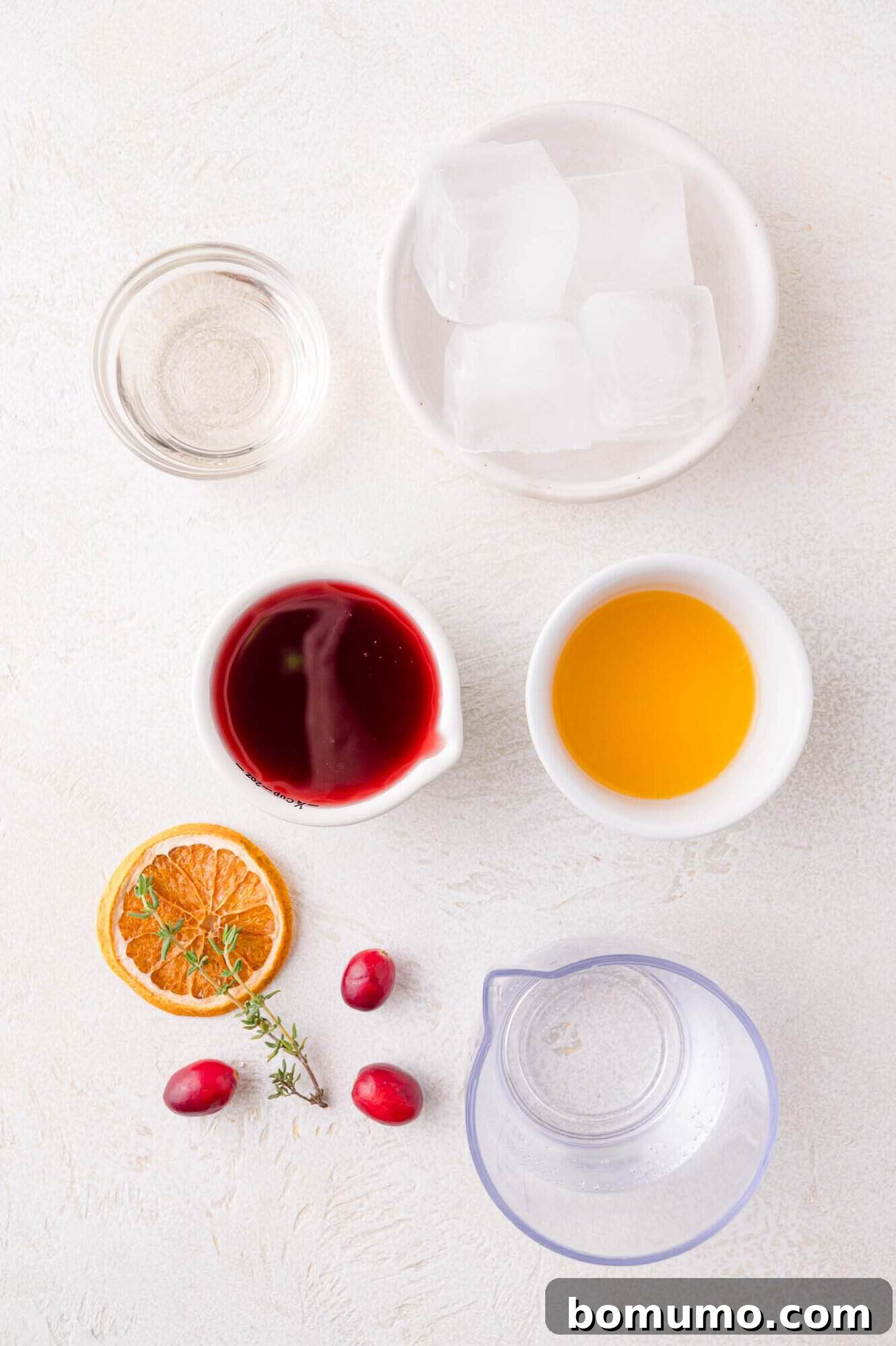 An overhead flat lay of the essential ingredients for the Cranberry Thyme Spritzer: ice cubes, cranberry juice, orange juice, simple syrup in a small pitcher, dried orange slices, and fresh cranberries and thyme for garnish, all arranged on a white surface.