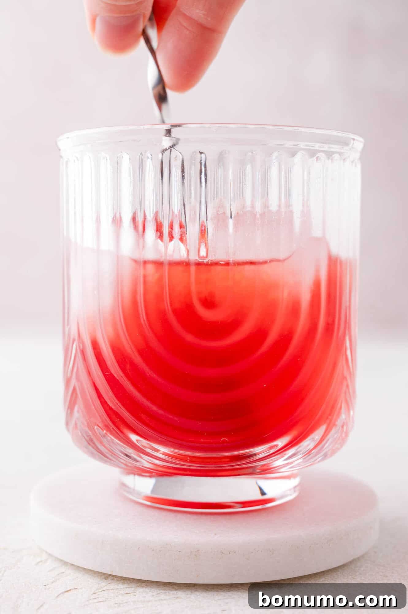 A hand gently stirs a ruby-red liquid in a clear, textured glass filled with ice, resting on a round white coaster. This captures the mixing of the juices and syrup.