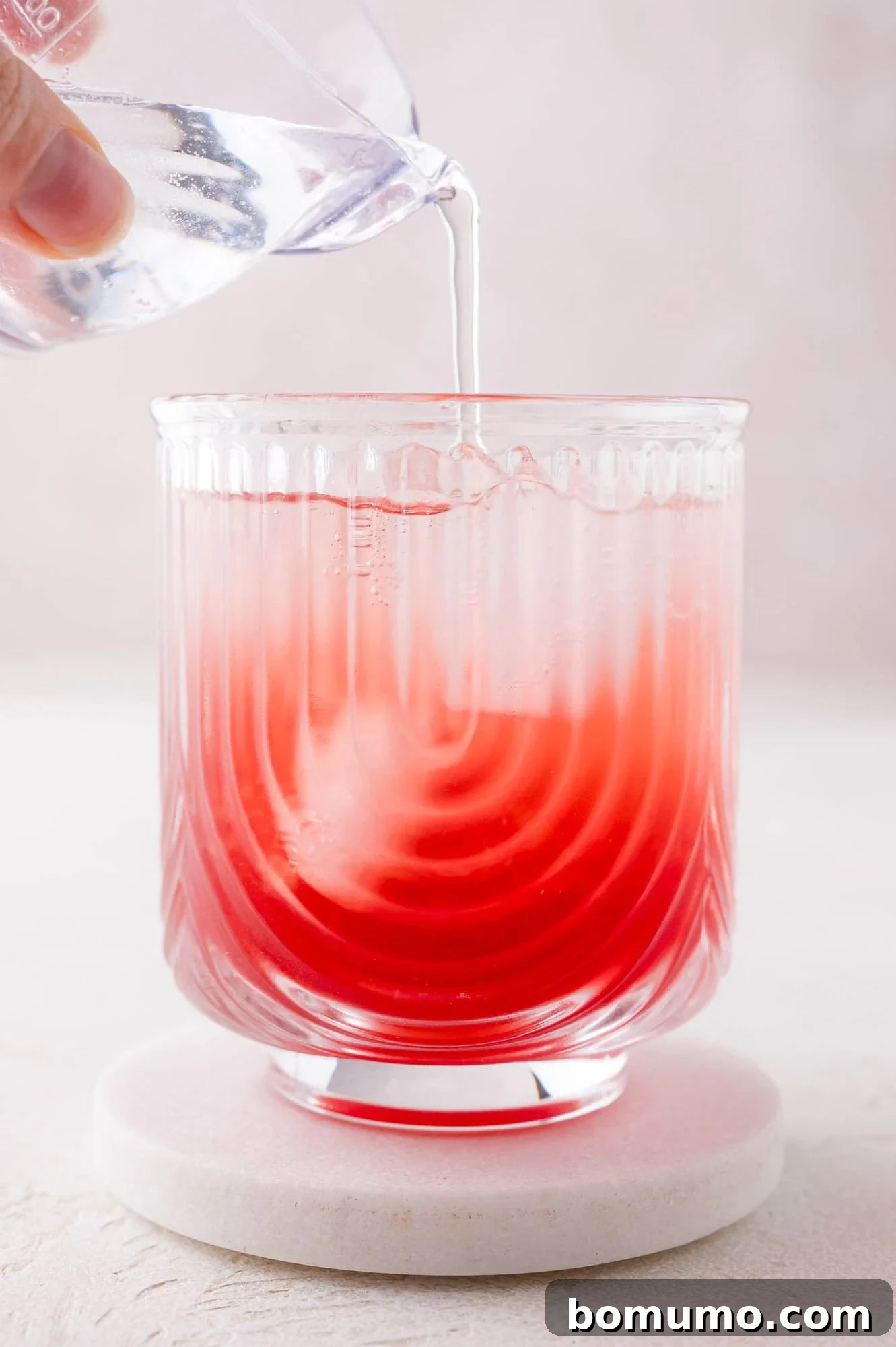 A clear liquid, sparkling water, is being carefully poured from a small glass into a larger textured glass containing the mixed cranberry and orange base, creating a beautiful swirling effect as the colors combine.