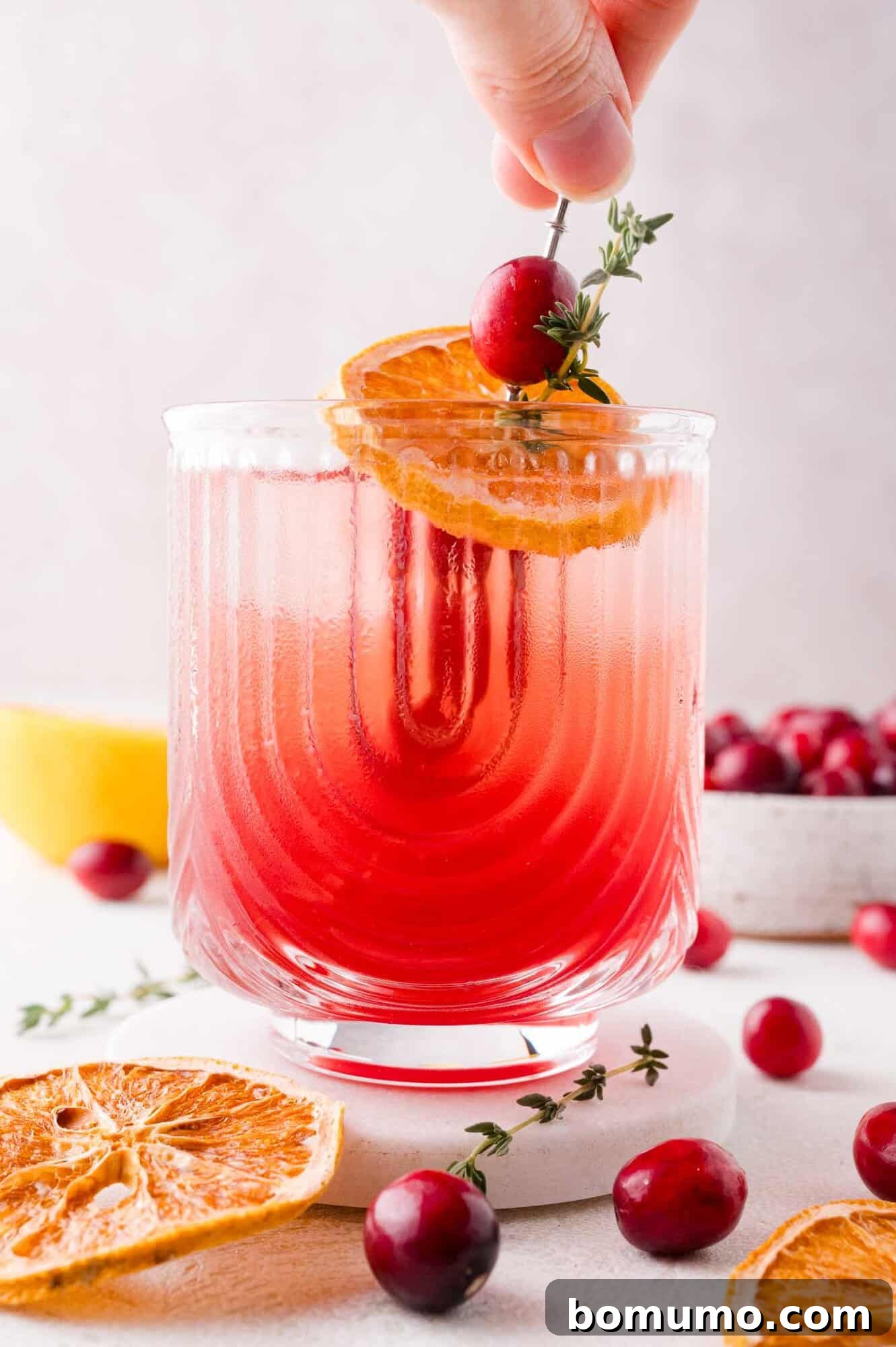 A hand meticulously garnishes a festive pink-red mocktail with a fresh cranberry, a dried orange slice, and a sprig of thyme. The elegant textured glass is surrounded by additional cranberries and orange slices on a white surface.