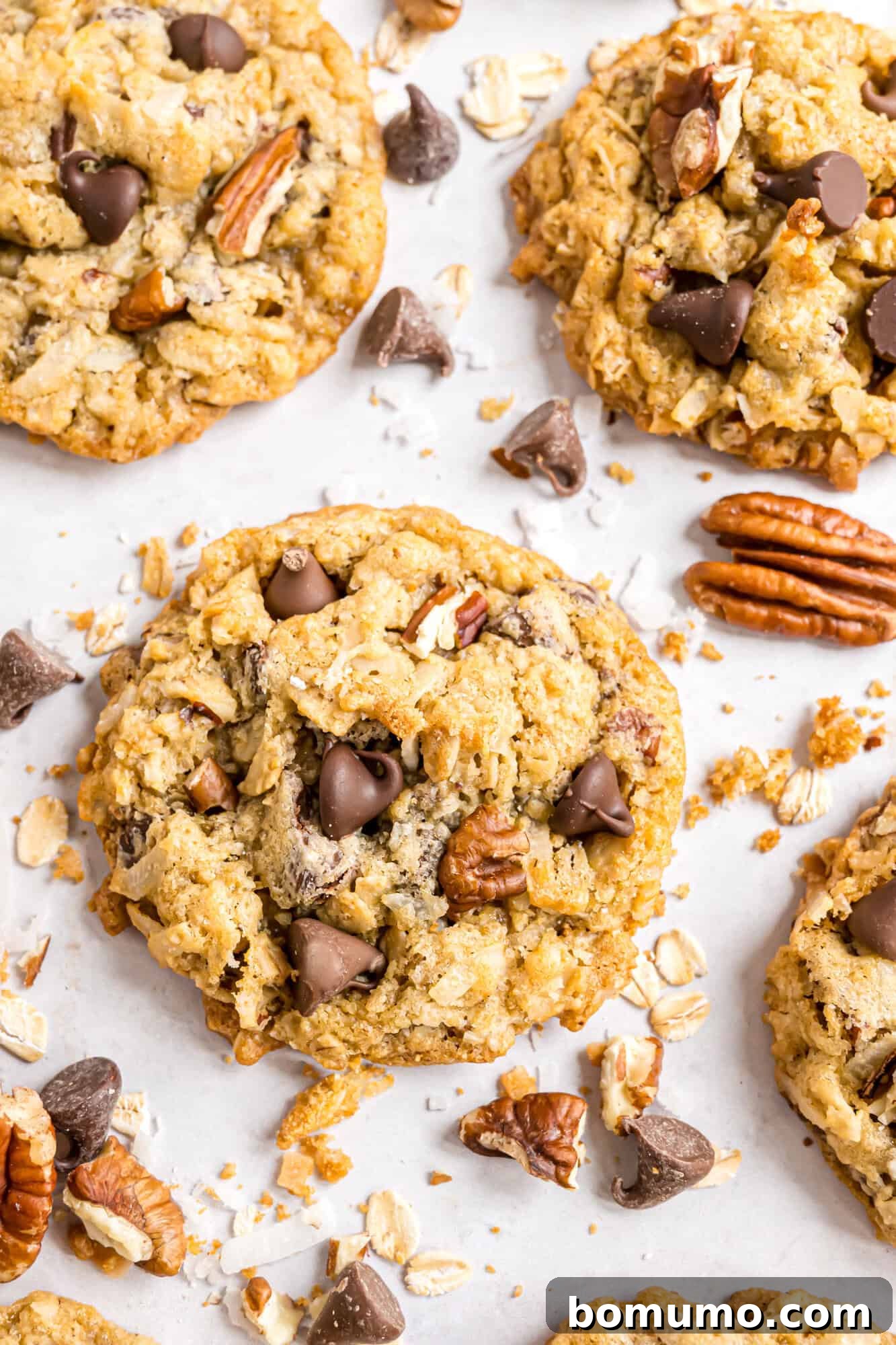 A close-up of cowboy cookies with chocolate chips and pecans, surrounded by scattered oats, chocolate chips, and pecan pieces on a white surface.
