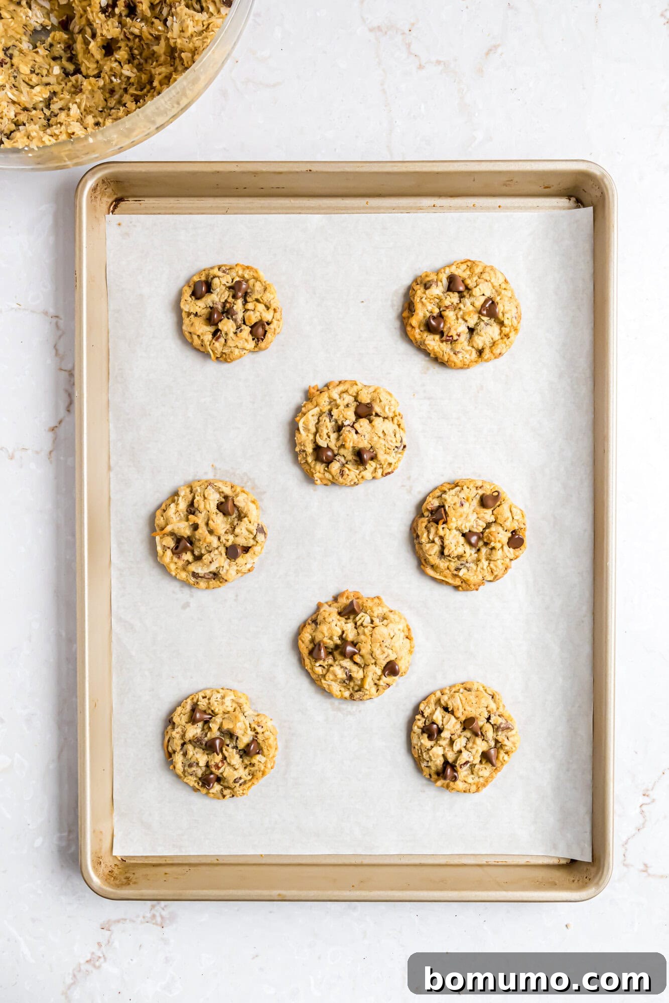 A baking sheet lined with parchment paper holds eight baked cowboy cookies, with more cookie dough visible in a mixing bowl at the top left corner.