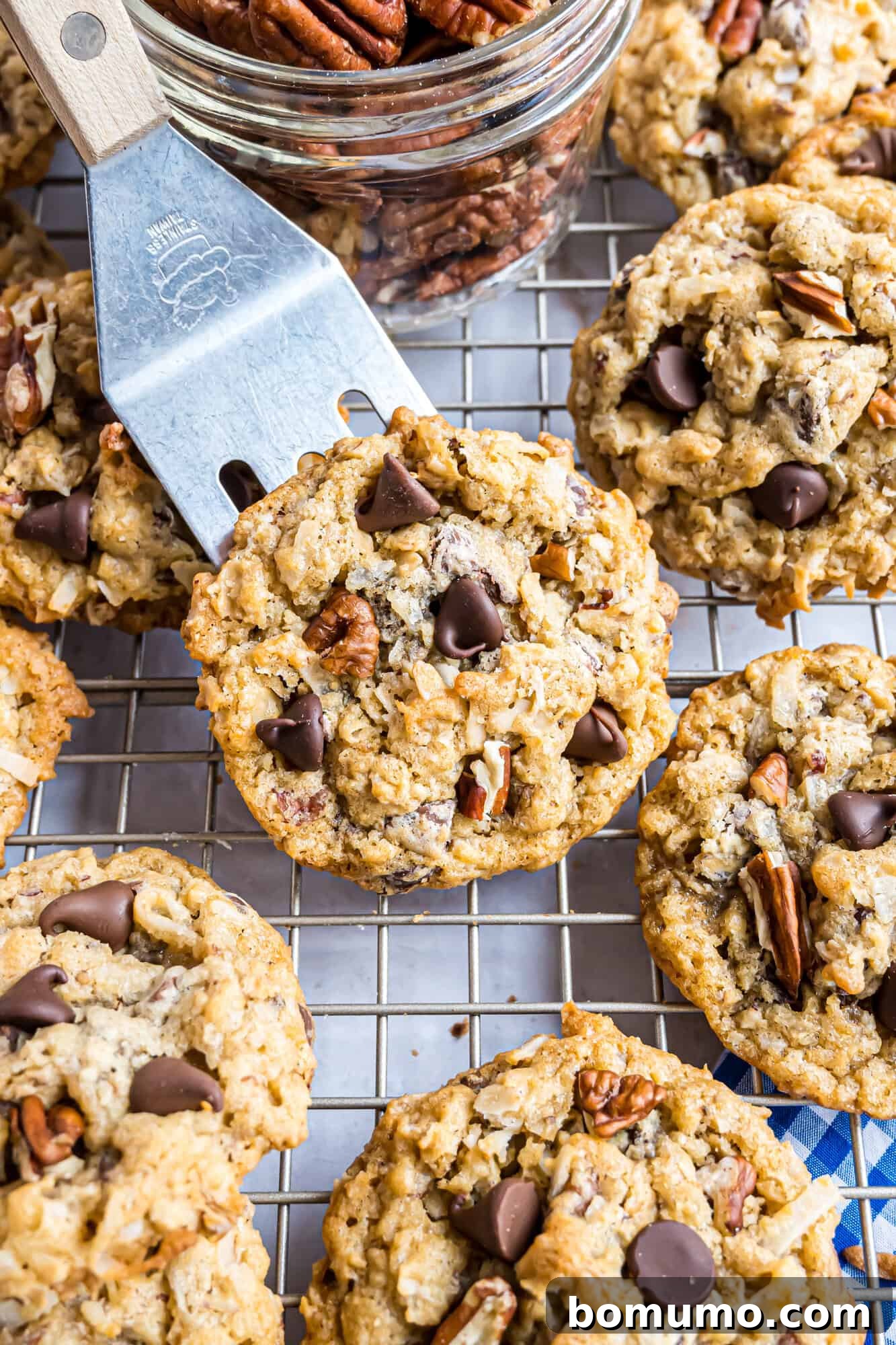 Cowboy cookies with chocolate chips and pecans on a cooling rack, with a spatula lifting one and a jar of pecans nearby.