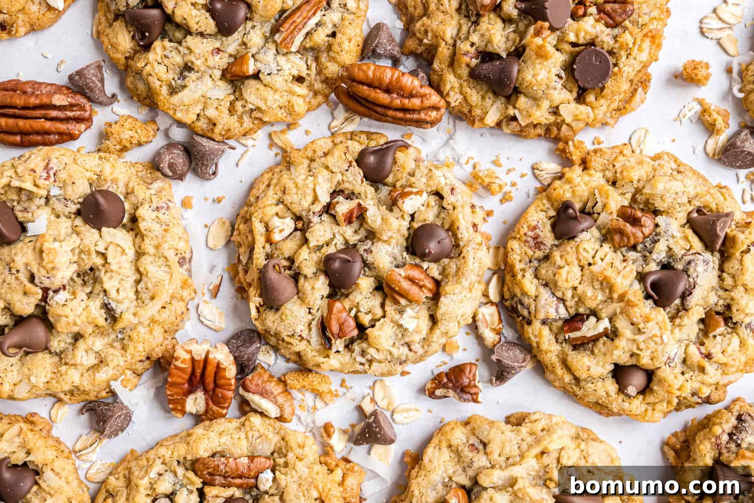 A close-up of oatmeal cookies topped with chocolate chips and pecans, with scattered pecans, oats, and chocolate pieces on a white surface.