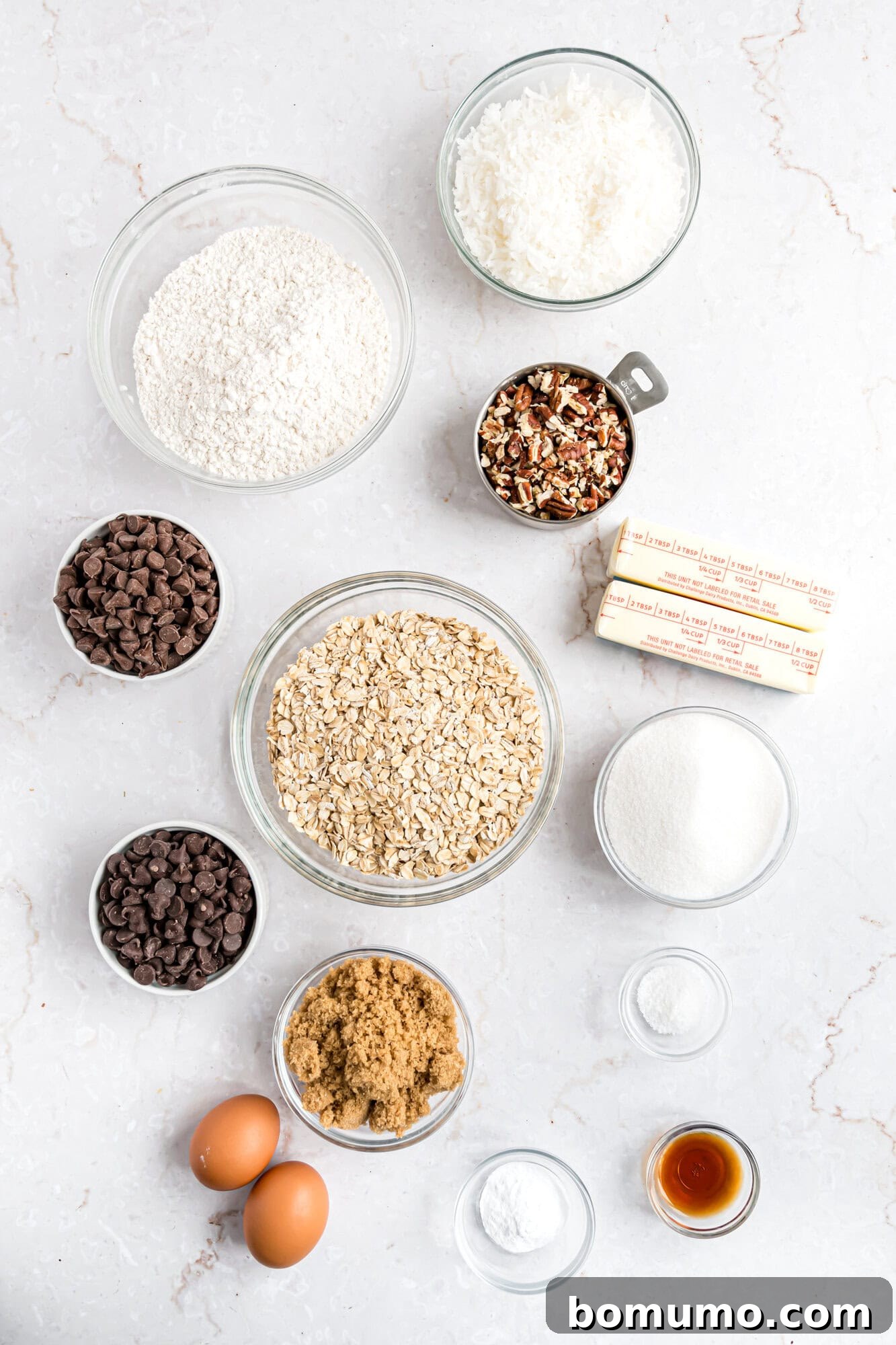Various baking ingredients in clear bowls on a white surface, including flour, oats, shredded coconut, pecans, chocolate chips, sugar, brown sugar, eggs, baking soda, vanilla, and butter.