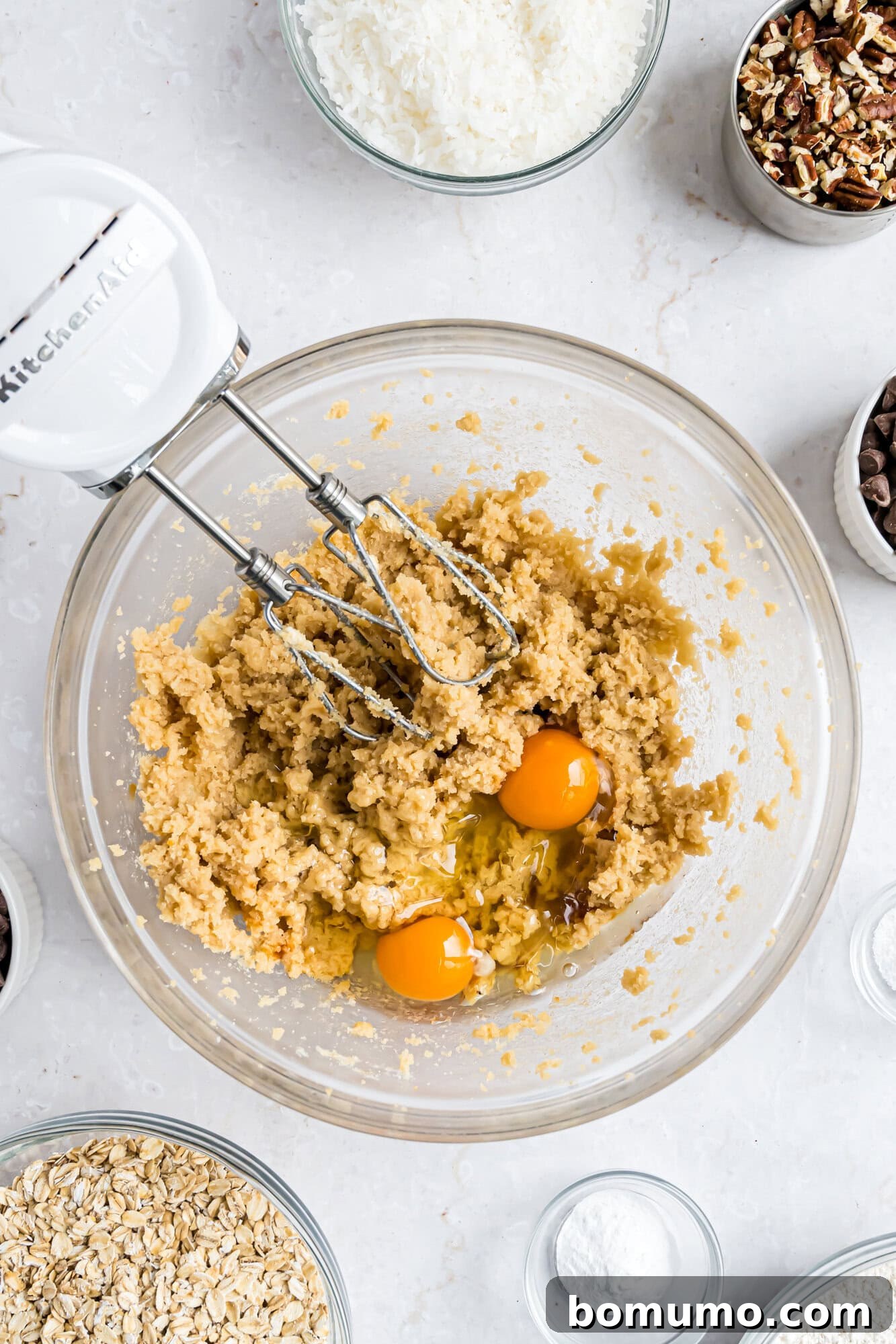 A glass bowl with cookie dough, two eggs, and a hand mixer surrounded by small bowls of oats, coconut, chocolate chips, pecans, and baking powder on a white surface.