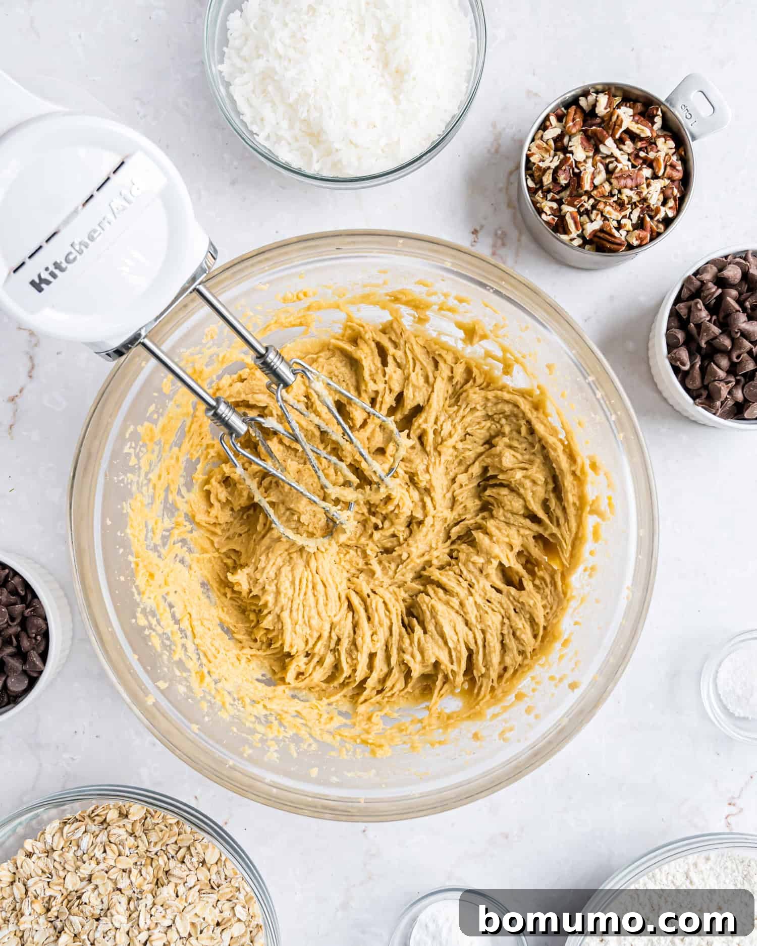 A stand mixer creaming cookie dough in a glass bowl, surrounded by bowls of oats, chocolate chips, chopped pecans, shredded coconut, and flour on a marble surface.