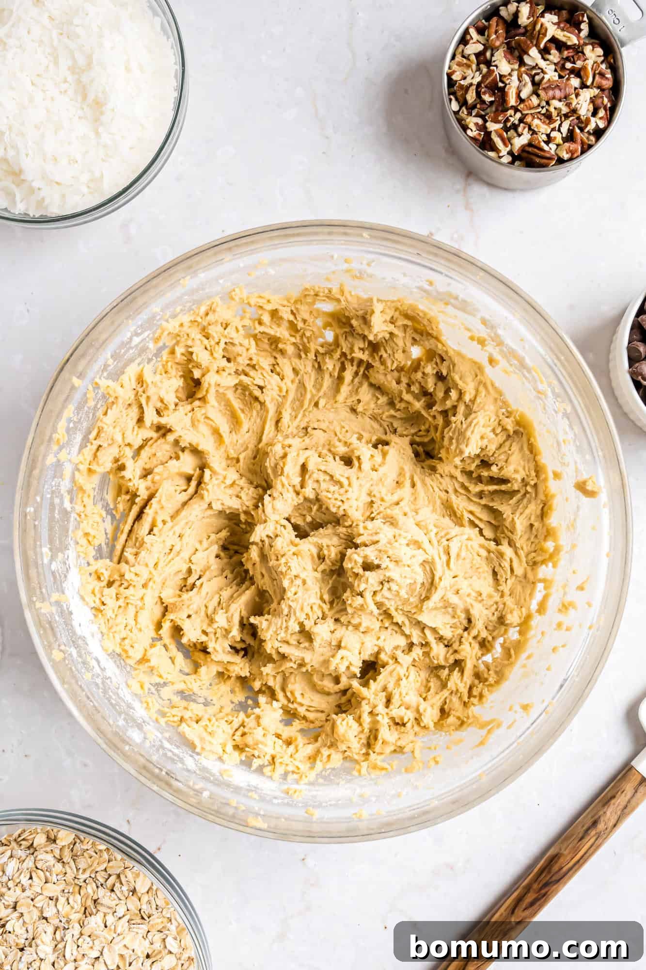 A glass bowl filled with cookie dough sits on a countertop, surrounded by bowls of chopped pecans, oats, shredded coconut, and chocolate chips.