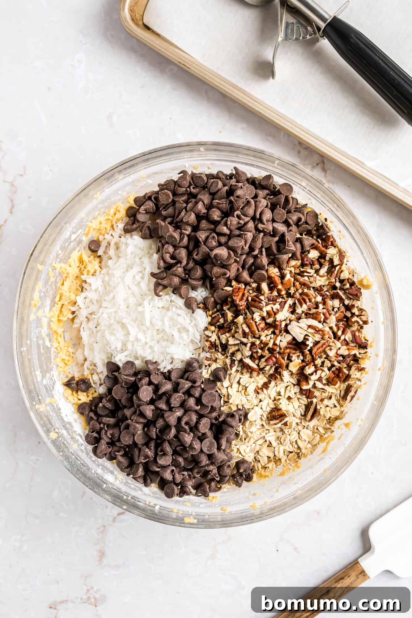 A glass bowl filled with chocolate chips, shredded coconut, chopped nuts, and oats, ready to be mixed; a baking tray and spatula are nearby.