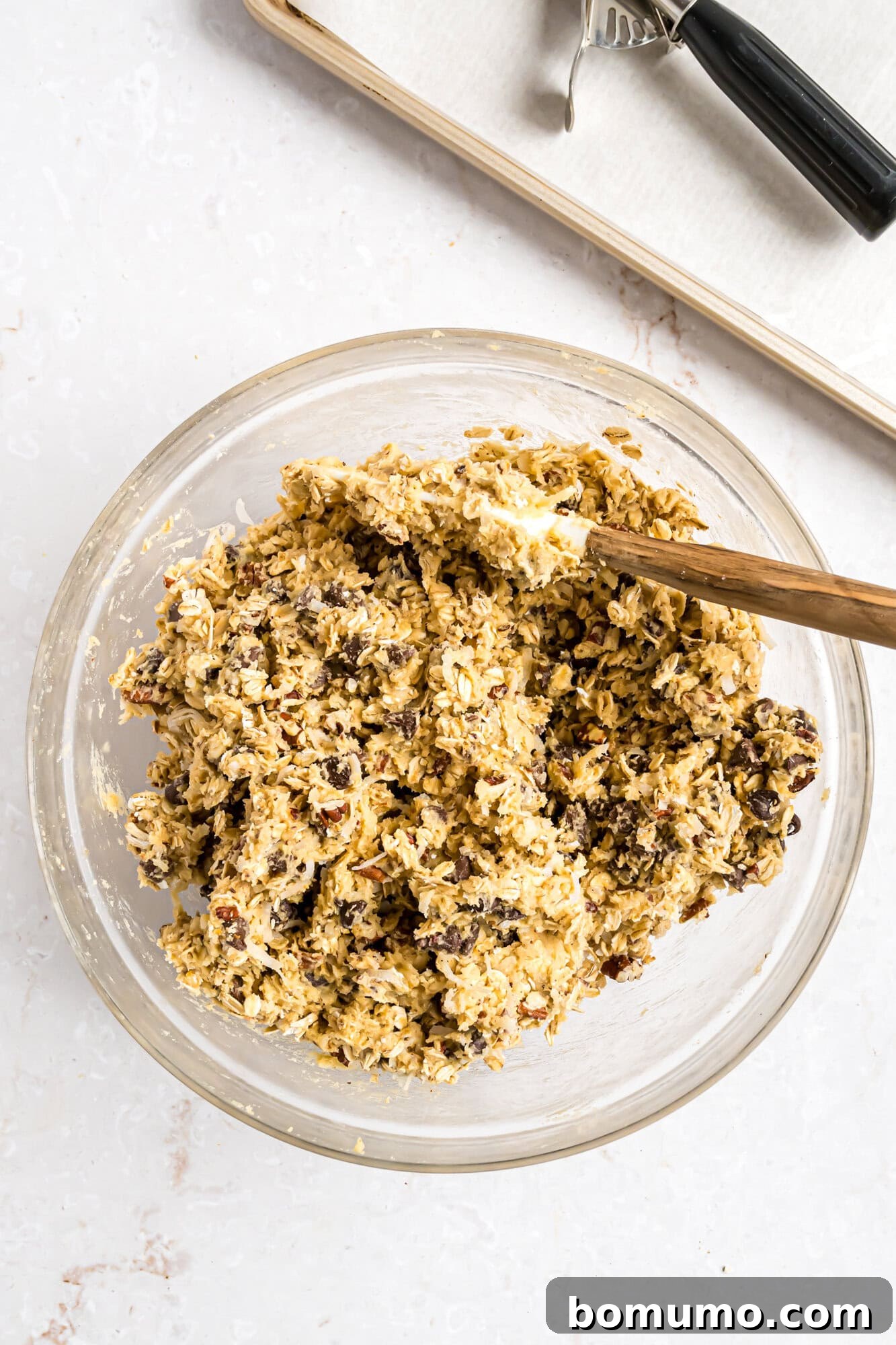 A glass bowl filled with cookie dough mixed with chocolate chips and nuts, with a wooden spoon resting inside. A baking sheet and cookie scoop are in the background.