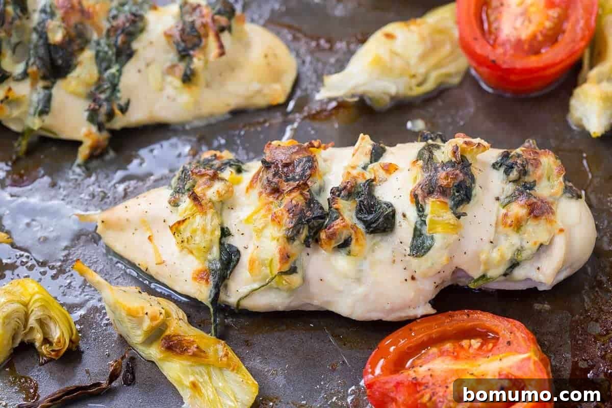 Close-up of golden-brown Hasselback chicken breast in a roasting pan, showcasing the melted cheese, spinach, and artichoke filling oozing from the deep cuts.