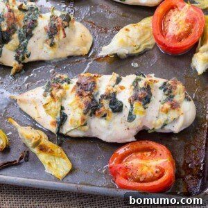 A sheet pan with Hasselback chicken, roasted tomatoes, and artichoke hearts. The chicken is stuffed with a creamy spinach and artichoke filling.