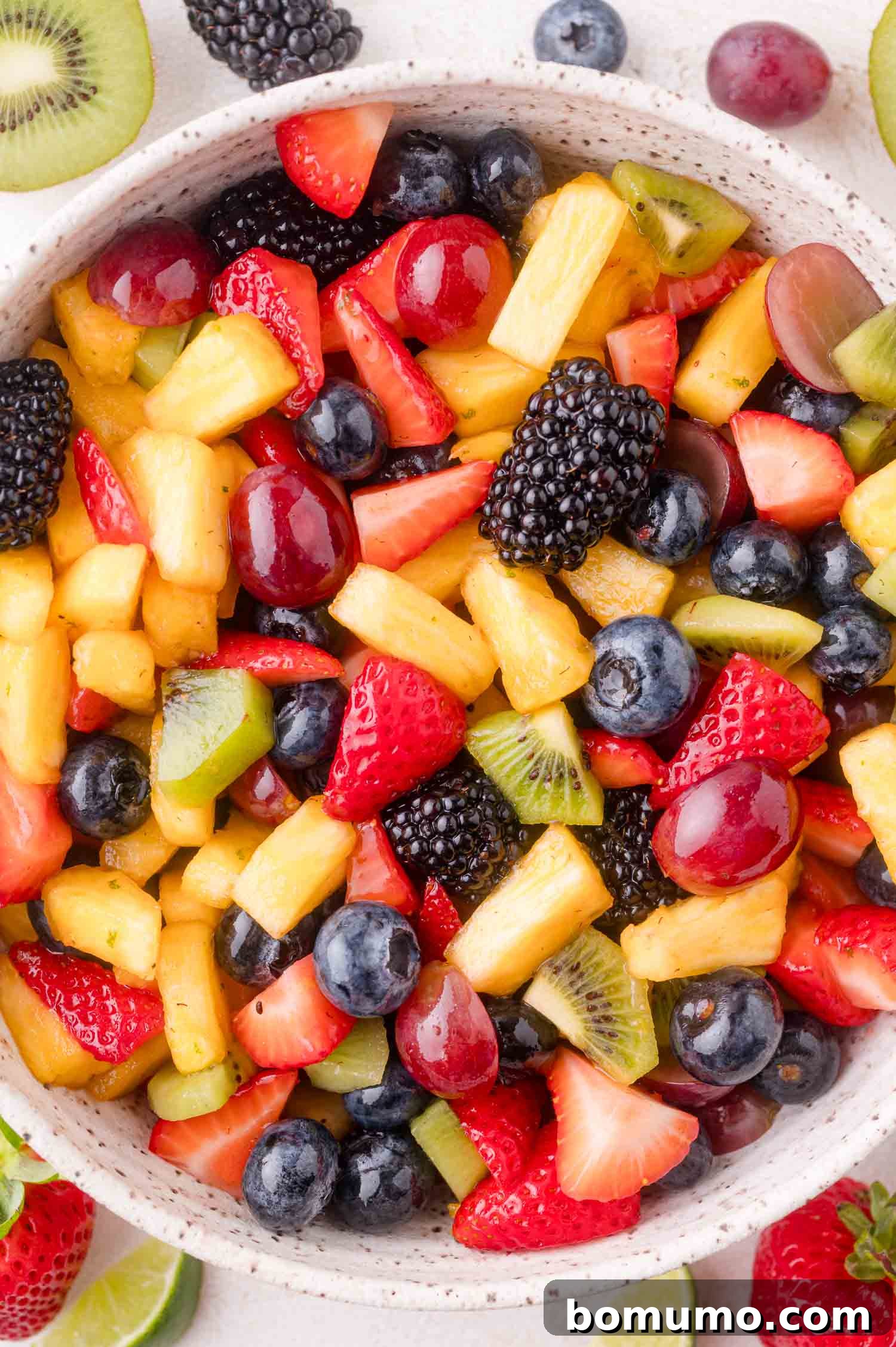 A beautifully arranged bowl brimming with a colorful assortment of fresh fruit, including bright red strawberries, vivid green kiwi, golden pineapple chunks, deep purple blackberries, succulent red grapes, and plump blueberries, all glistening with a light dressing.