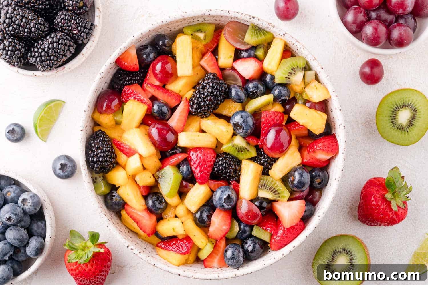 A vibrant bowl of mixed fruit salad featuring golden pineapple, bright red strawberries, plump blueberries, dark blackberries, sweet green grapes, and tangy kiwi, elegantly arranged on a white surface with small bowls of additional berries and fruit slices.
