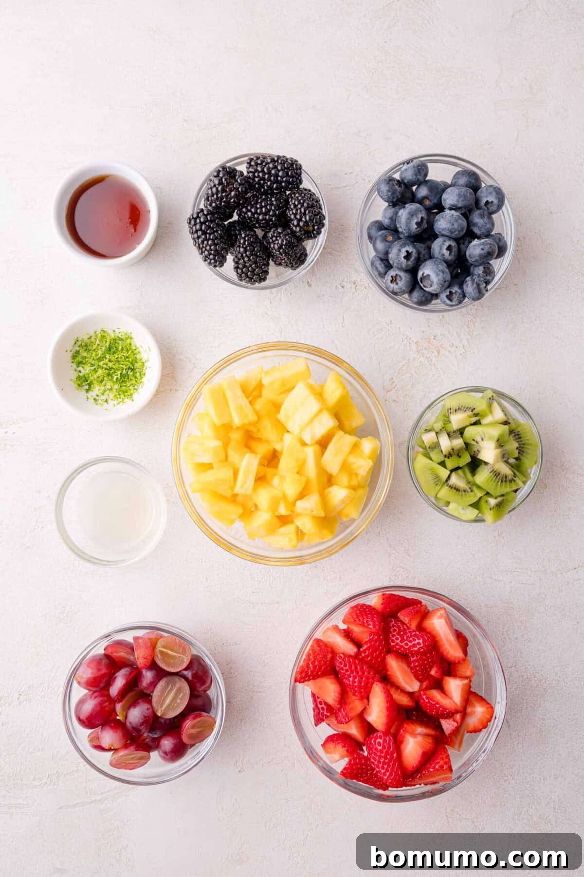 A collection of bowls filled with prepared fruit components for the salad, including diced pineapple, hulled and quartered strawberries, halved red grapes, blueberries, blackberries, and diced kiwi, alongside small dishes holding fresh lime juice, lime zest, and honey, all arranged on a pristine white surface.