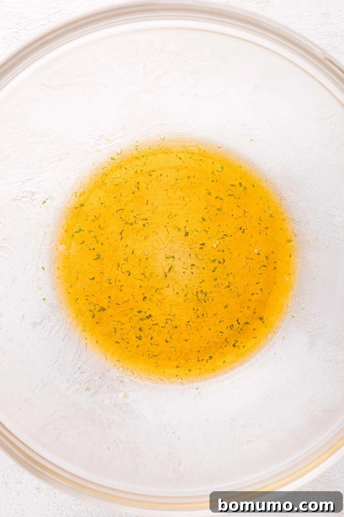 A close-up view of the freshly prepared honey-lime dressing in a clear glass bowl, featuring a smooth yellow liquid infused with fine green zest particles, signifying its readiness to coat the fruit.