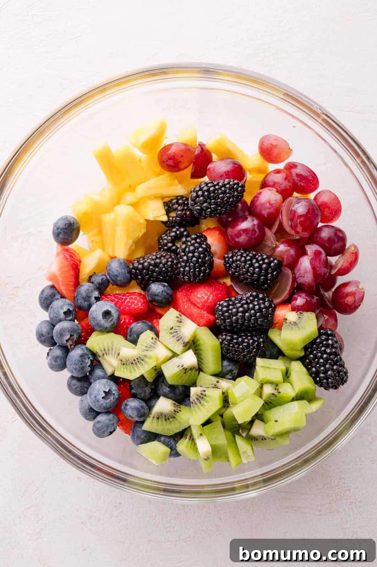 A large glass bowl filled with a delightful medley of freshly sliced kiwi, plump blackberries, juicy blueberries, vibrant strawberries, golden pineapple chunks, and sweet red grapes, all awaiting the final dressing.