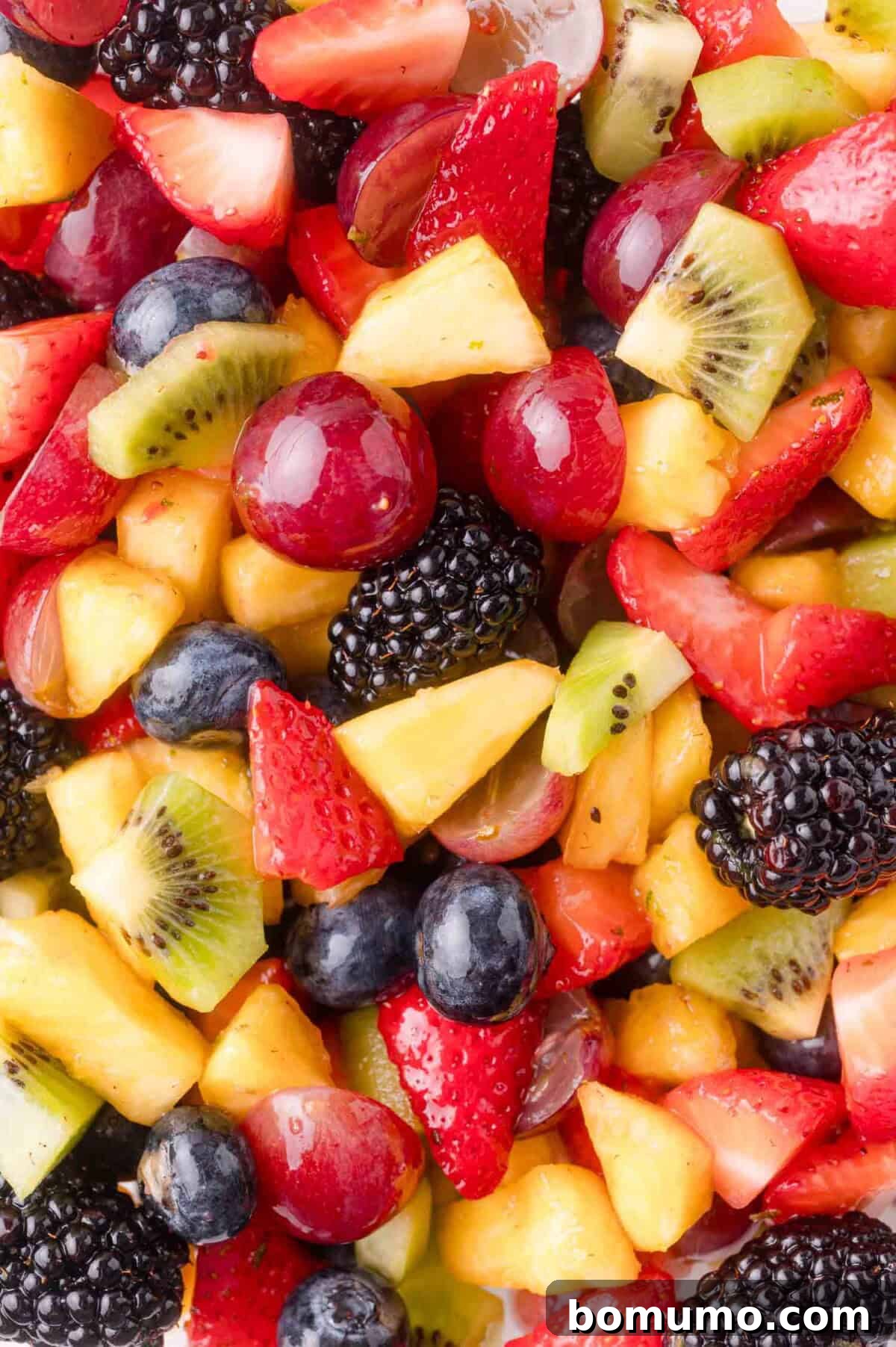 A vivid close-up shot of the mixed fruit salad, showcasing the delightful blend of red strawberries, blue blueberries, dark blackberries, green kiwi, yellow pineapple, and red grapes, all glistening with a fresh, light dressing.