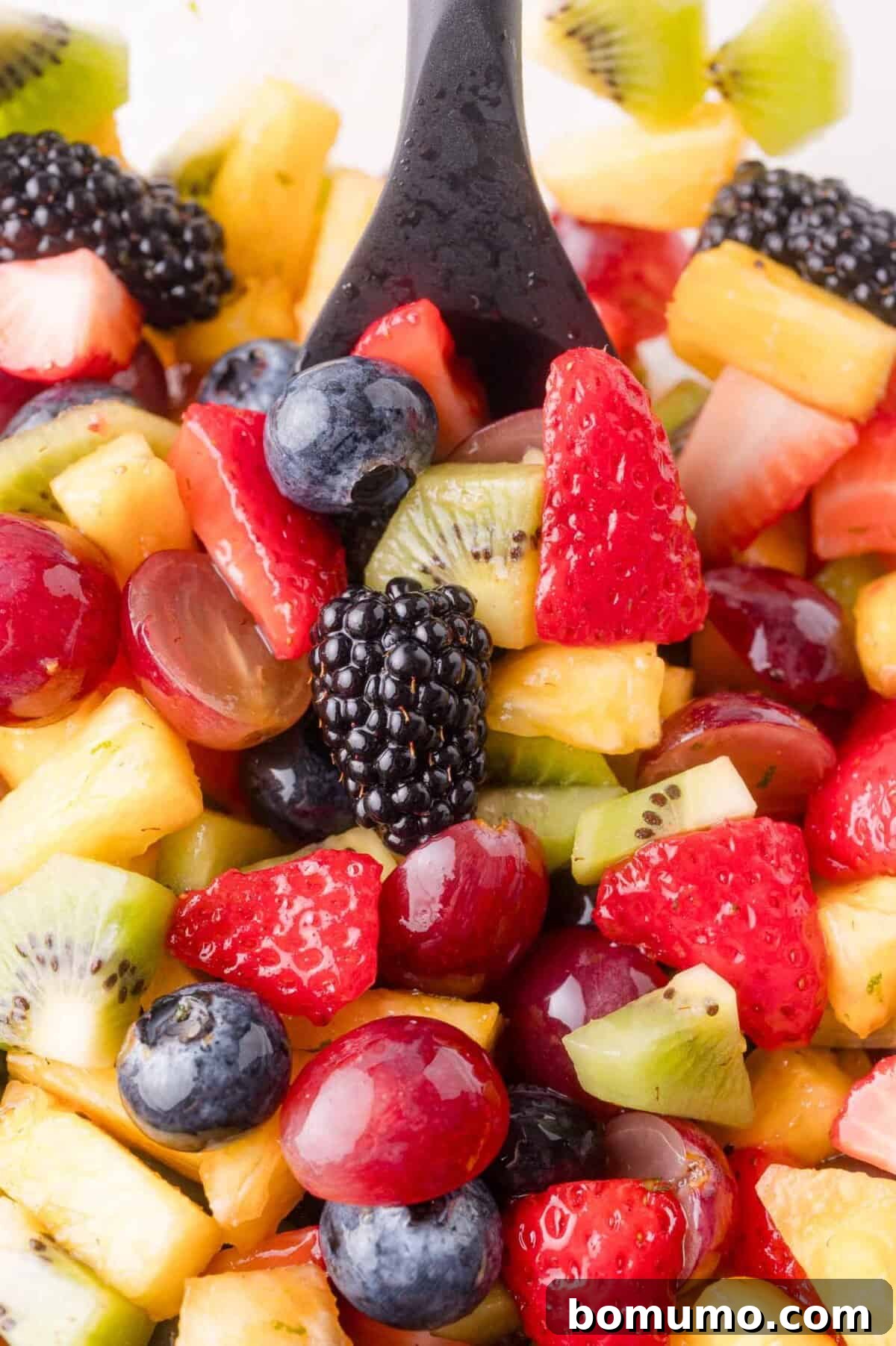 A close-up view of a refreshing fruit salad featuring a medley of strawberries, kiwi, pineapple, grapes, blackberries, and blueberries, with a black serving spoon perfectly positioned within the colorful mix, ready to be enjoyed.
