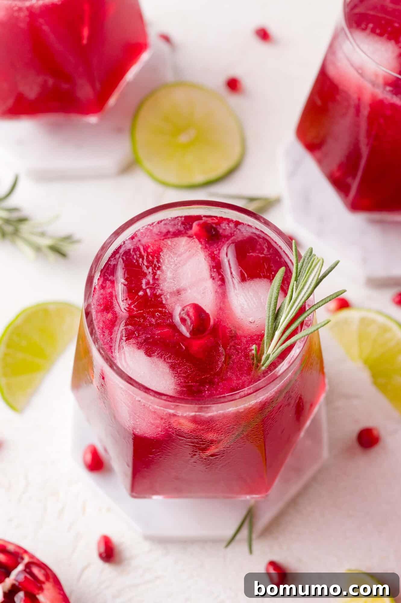 A vibrant red pomegranate mocktail in a highball glass filled with ice, elegantly garnished with a fresh sprig of rosemary. The drink is surrounded by glistening lime slices and ruby red pomegranate seeds on a clean white surface, highlighting its festive appeal and refreshing nature.