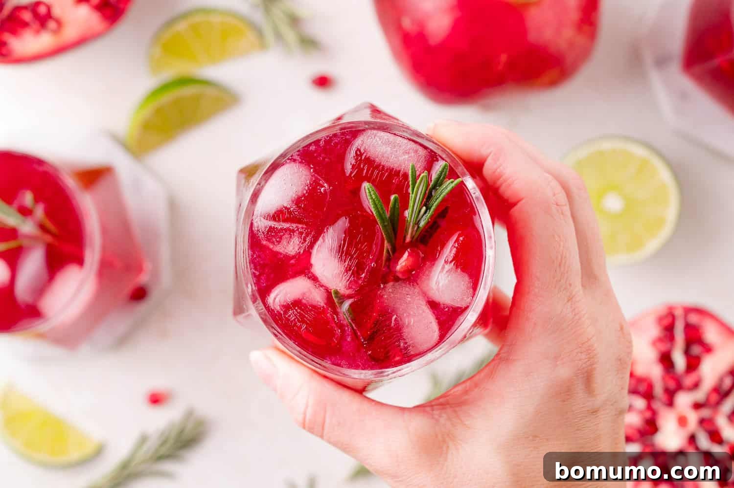 A hand elegantly holds a highball glass of pomegranate spritzer, beautifully garnished with a sprig of rosemary and a scattering of pomegranate seeds. The background shows vibrant lime slices and additional pomegranate arils on a pristine white table, emphasizing its freshness and appeal.
