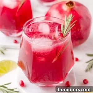 A glass of red pomegranate spritzer with ice and a rosemary sprig garnish sits on a coaster, with a whole pomegranate and lime slices in the background.