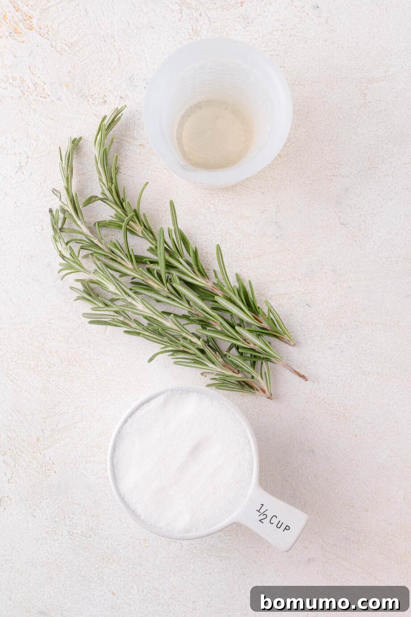 A simple setup for making rosemary simple syrup, showing a plastic cup of water, fresh rosemary sprig, and a measuring cup of granulated sugar on a light background, ready for infusion.