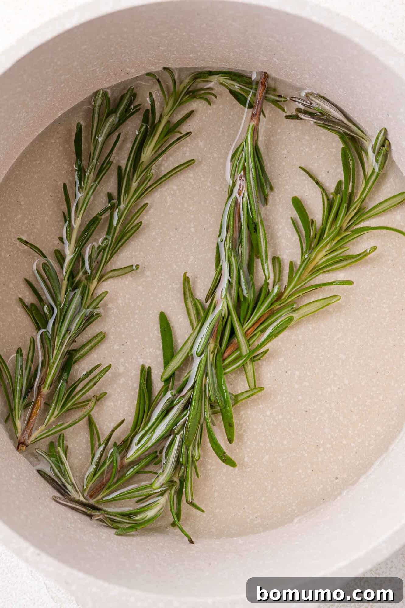 Two sprigs of vibrant green fresh rosemary gently steeping in clear simple syrup within a light-colored saucepan, infusing the liquid with aromatic herbal notes.
