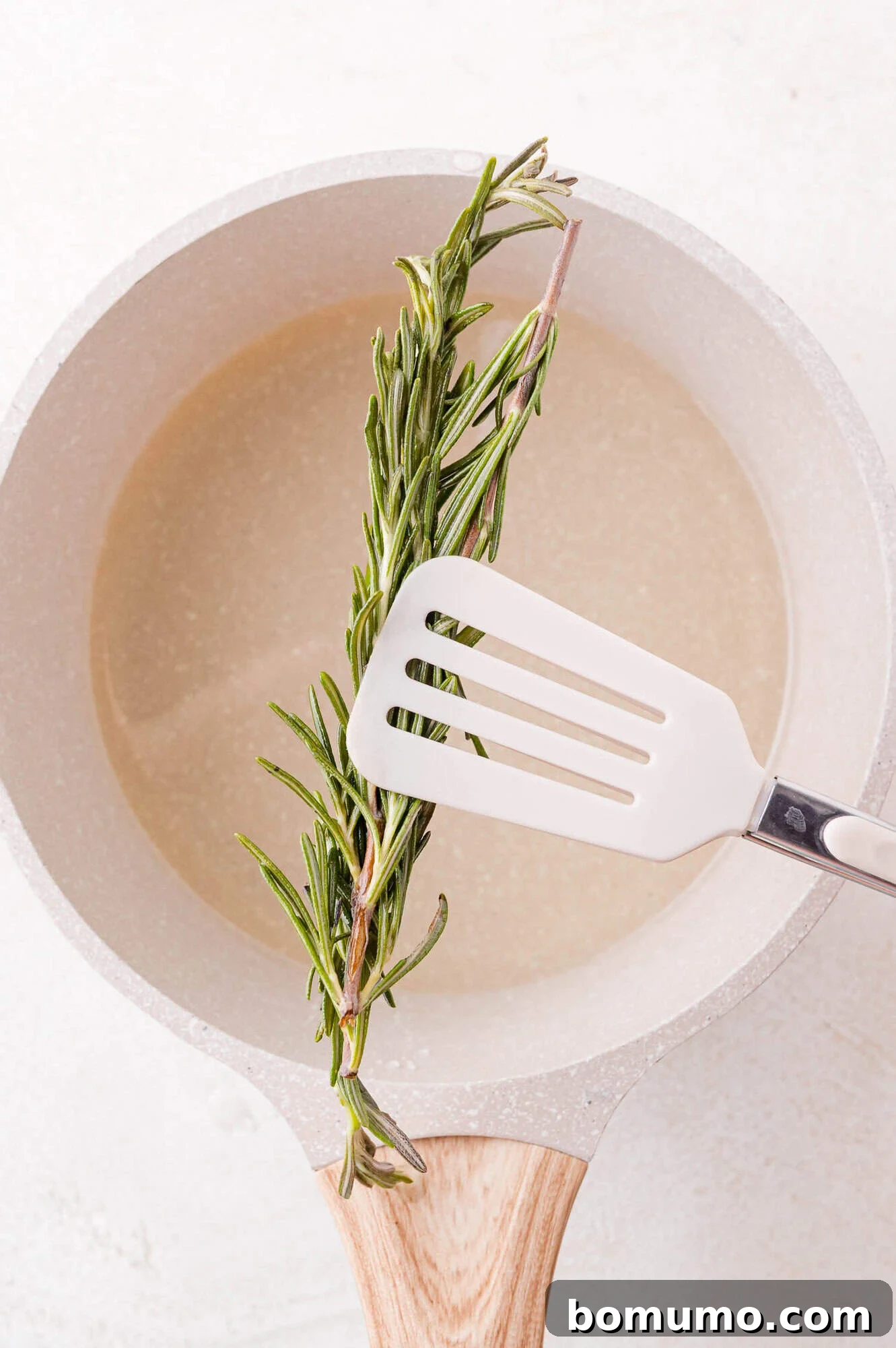 A white spatula carefully lifts a sprig of fresh rosemary from a saucepan filled with simple syrup, indicating the completion of the steeping process.