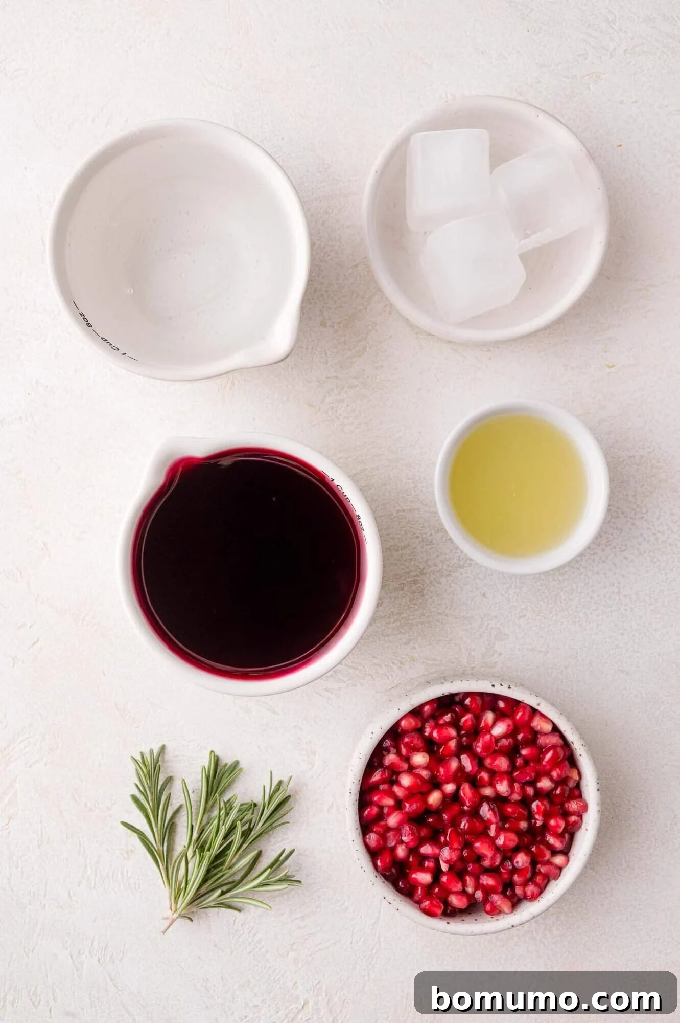 A collection of fresh ingredients for pomegranate spritzers: bowls containing vibrant pomegranate juice, ruby-red pomegranate seeds, freshly squeezed lime juice, clear ice cubes, and a fragrant rosemary sprig, all neatly arranged on a white surface.