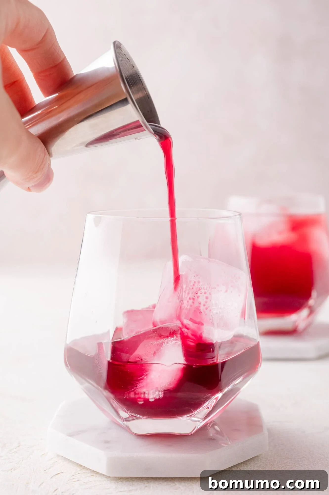 A hand expertly pours rich red pomegranate juice from a jigger into a highball glass filled with ice cubes, with a second prepared glass blurred in the background, illustrating the drink assembly.