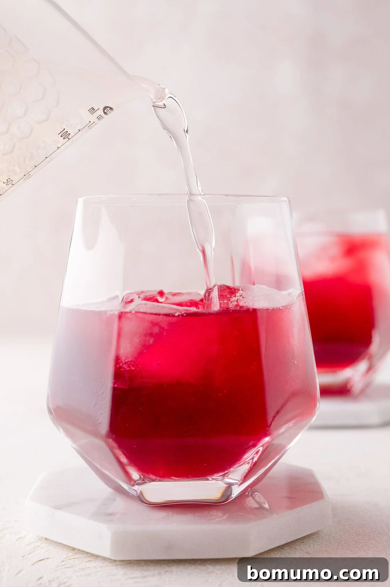 A close-up view of a highball glass containing pomegranate juice and ice cubes, as sparkling water is poured in, creating effervescent bubbles. The glass rests on a white coaster, with another similar glass softly blurred in the background.