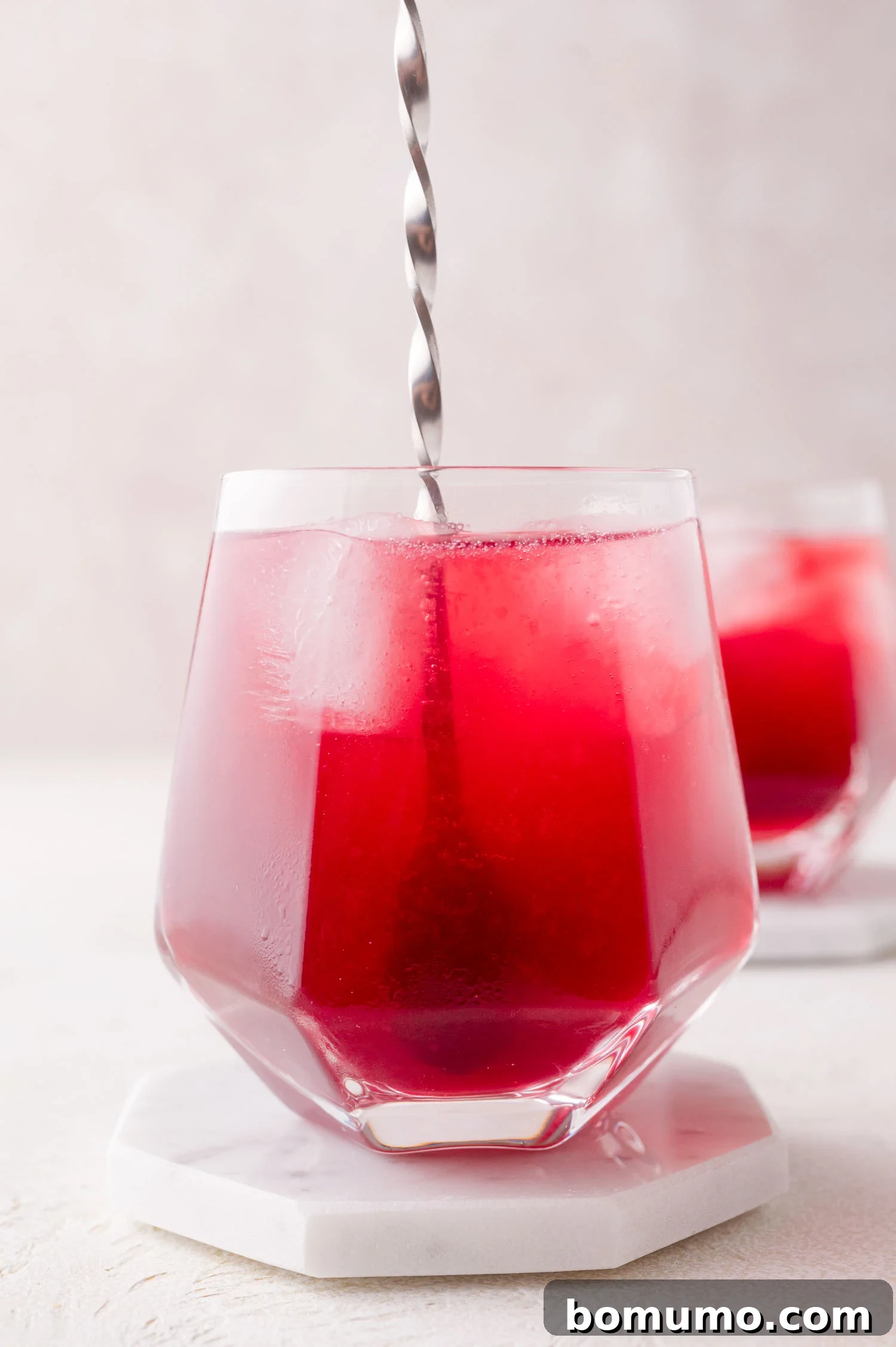 A ruby-red pomegranate spritzer in a highball glass is gently stirred with a slender metal spoon, its effervescent bubbles catching the light. The glass is placed on a stylish white hexagonal coaster, with another glass in the soft background.