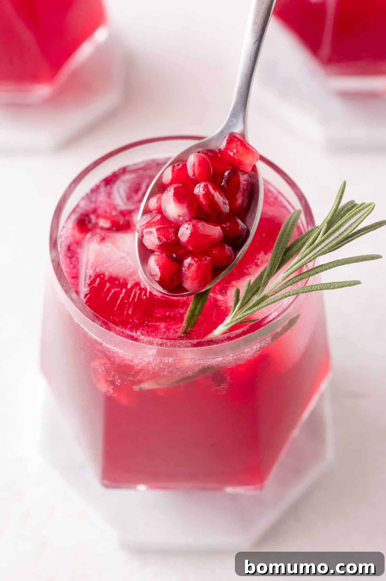 A beautifully garnished glass of red pomegranate spritzer, featuring ice, a fresh rosemary sprig, and a spoonful of glistening pomegranate seeds held just above the drink, ready to be dropped in for the final touch.