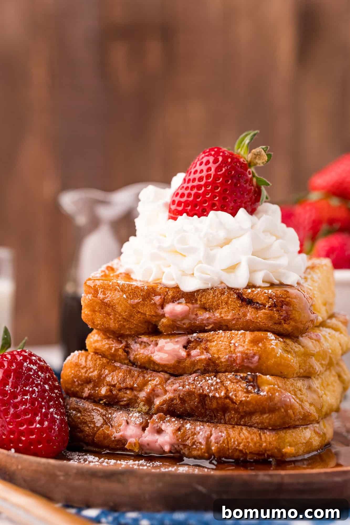 Stack of stuffed french toast topped with fresh strawberries and a dollop of whipped cream, drizzled with maple syrup.