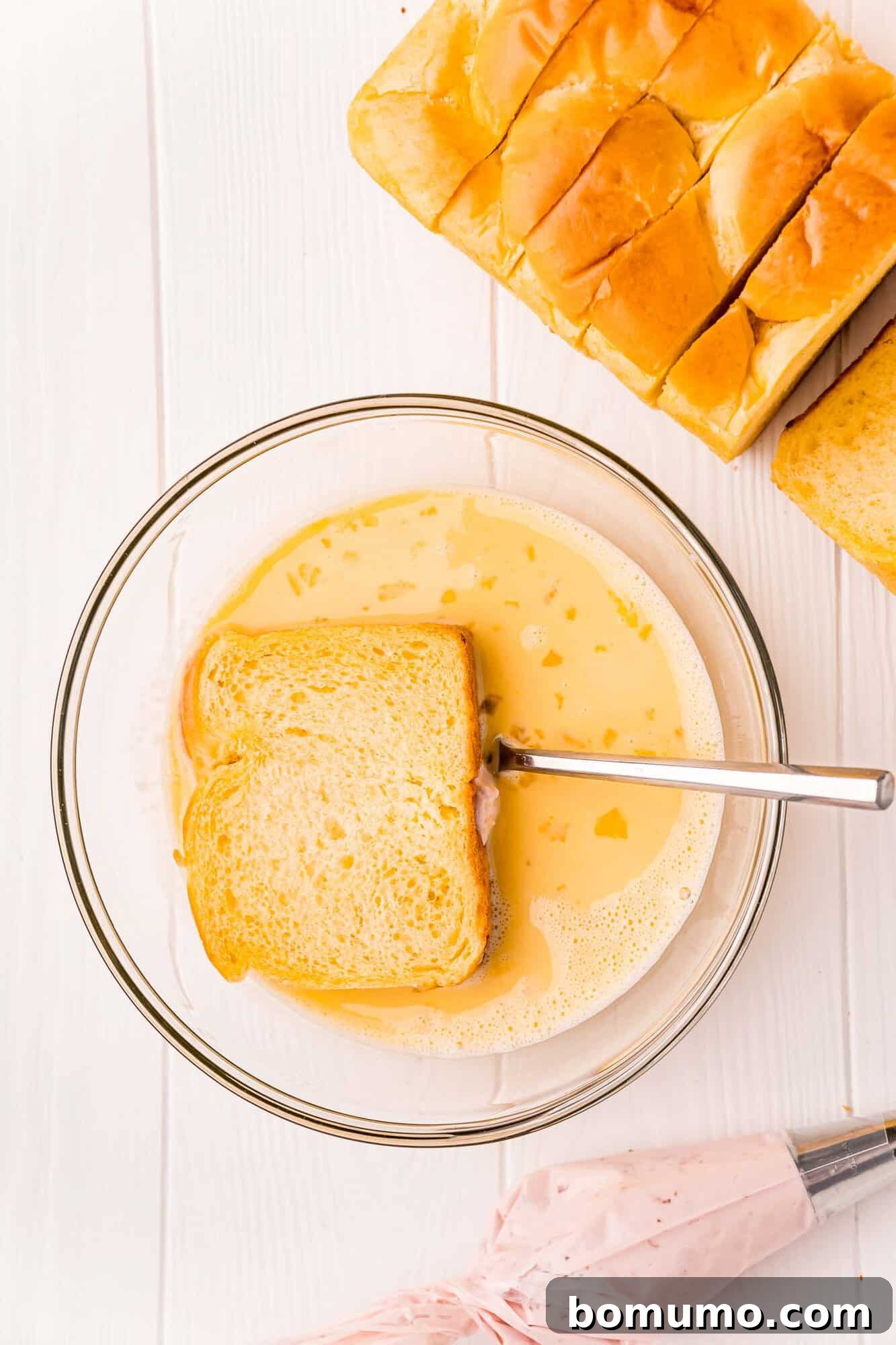 A slice of stuffed brioche bread being carefully dipped into the creamy egg mixture in a wide, shallow bowl.