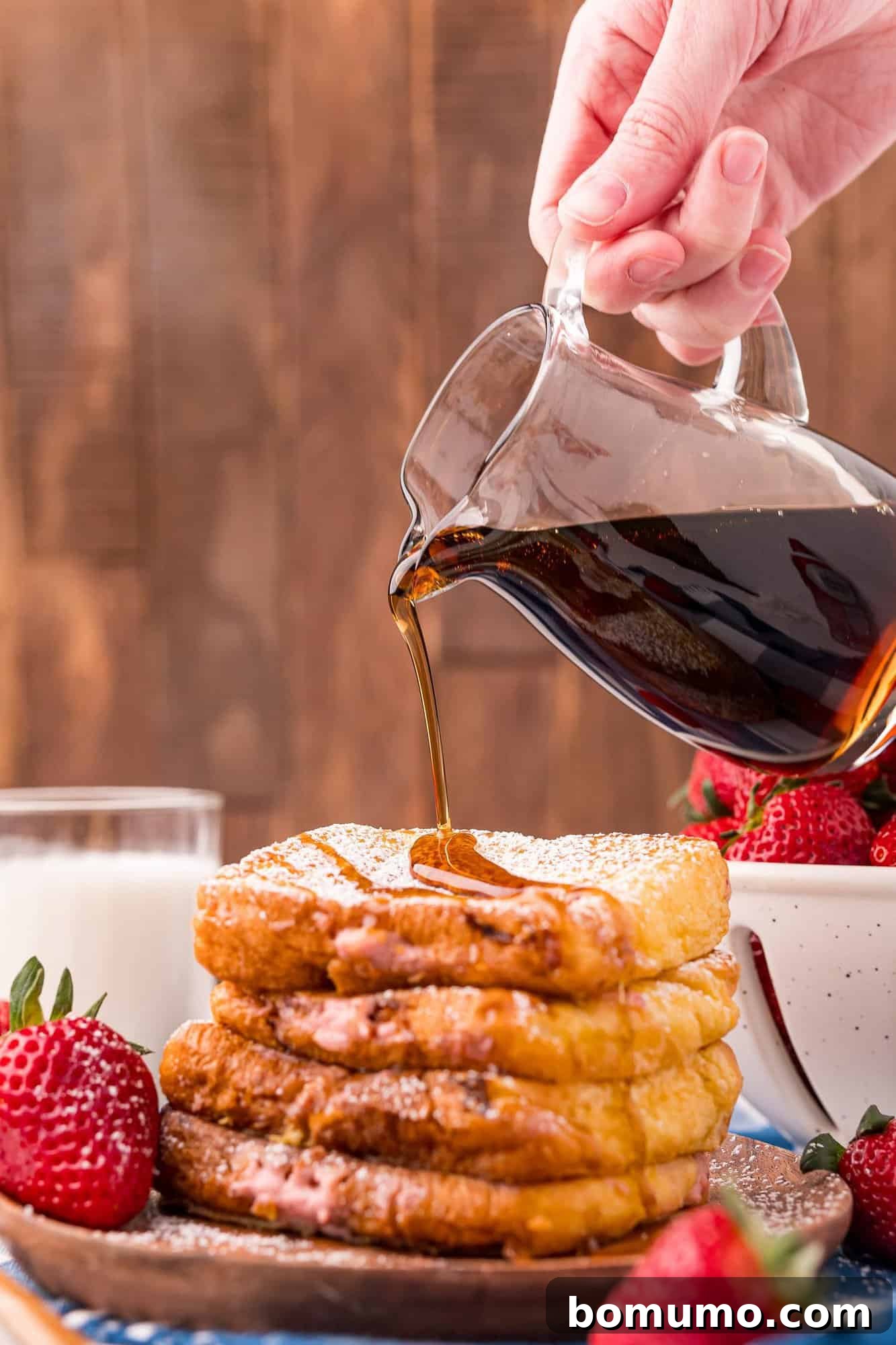 Rich maple syrup being poured generously over a beautifully stacked plate of strawberry cream cheese stuffed French toast.