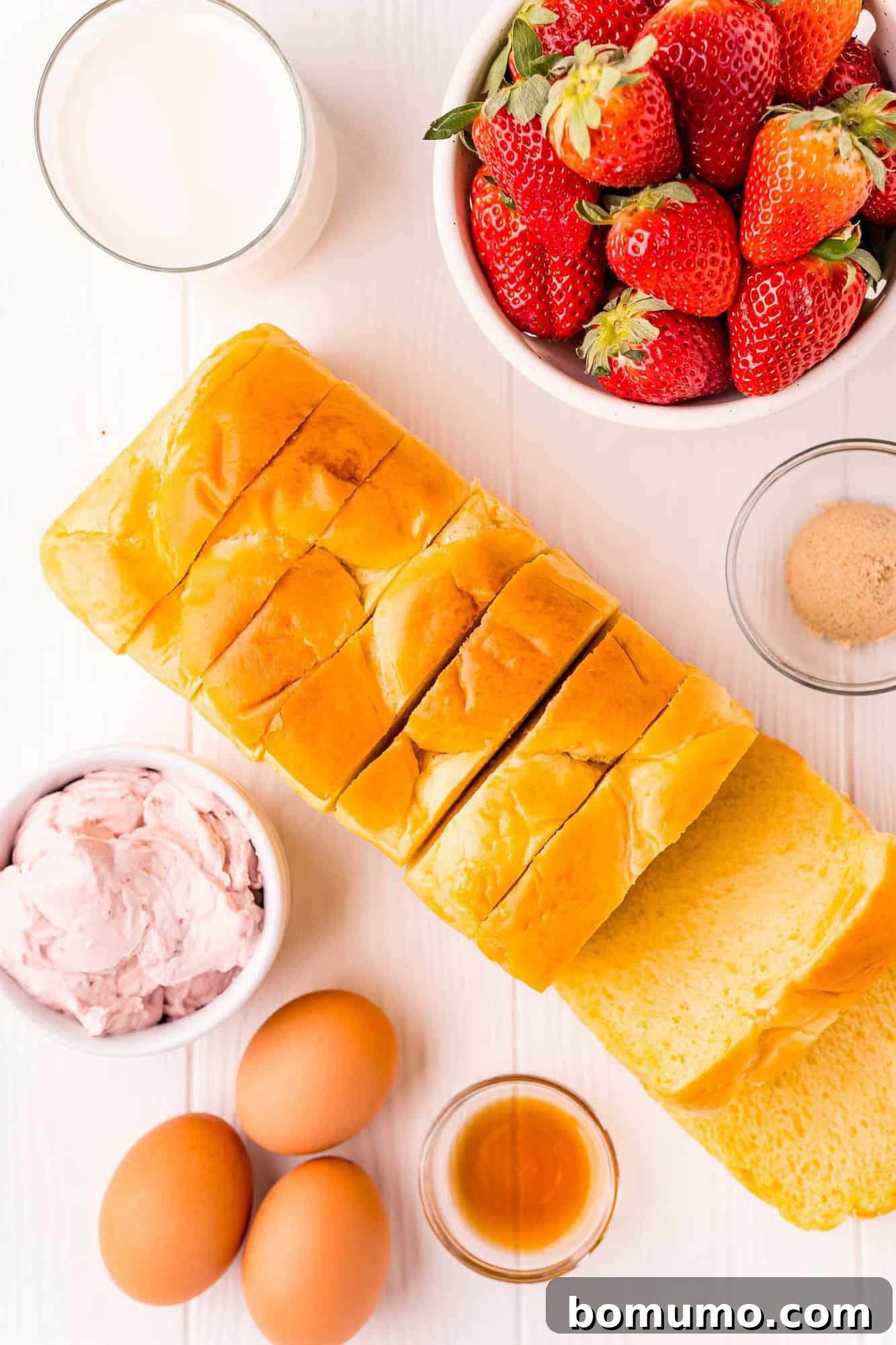 Overhead view of fresh ingredients needed for making strawberry cream cheese stuffed french toast, including brioche bread, eggs, milk, strawberry cream cheese, vanilla extract, and brown sugar.