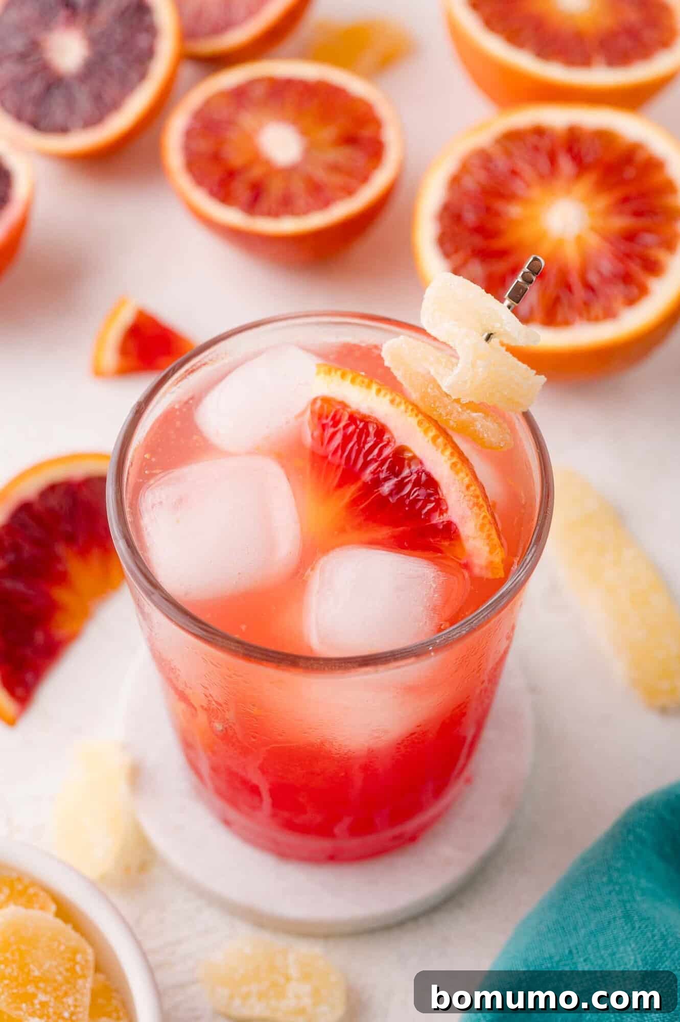 Crimson Sunset Sip 2 A vibrant blood orange mocktail, garnished with a fresh blood orange slice and candied ginger, sits invitingly on ice, surrounded by halved blood oranges and ginger pieces on a bright surface.