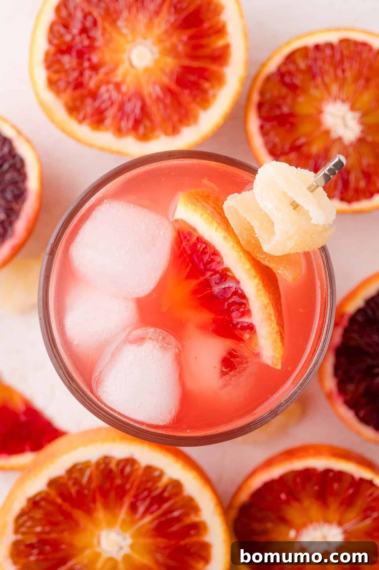Crimson Sunset Sip 11 A beautifully presented glass of blood orange mocktail with ice, garnished with a vibrant blood orange slice and a twist of candied ginger, artfully surrounded by halved blood oranges.