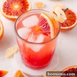 Crimson Sunset Sip 12 A sparkling blood orange mocktail, beautifully presented with ice, a blood orange wedge, and candied ginger, set on a white coaster amidst sliced blood oranges and ginger.
