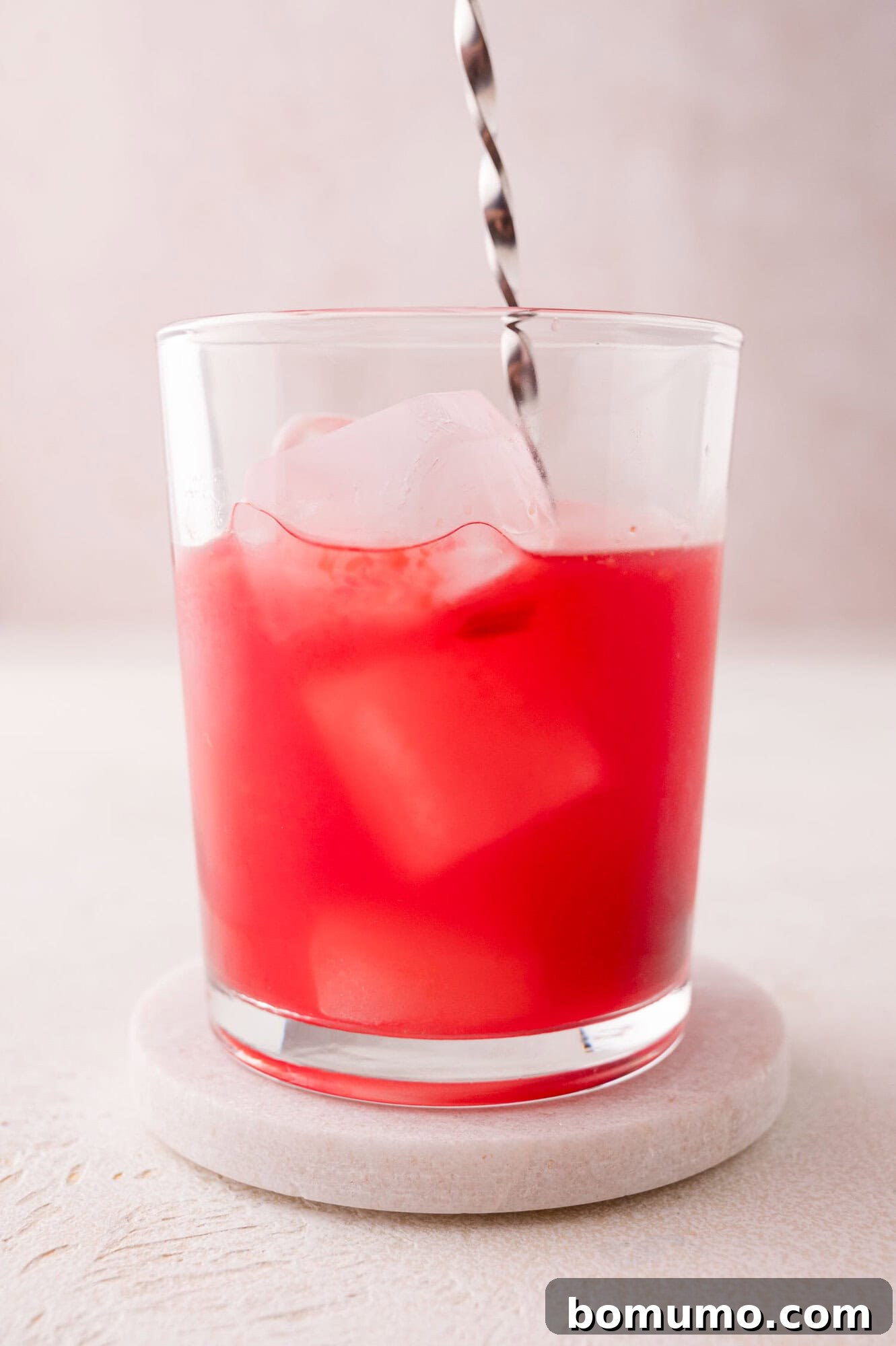 Crimson Sunset Sip 7 A clear glass filled with a bright red liquid and ice cubes, being stirred with a metal spoon to combine ingredients.
