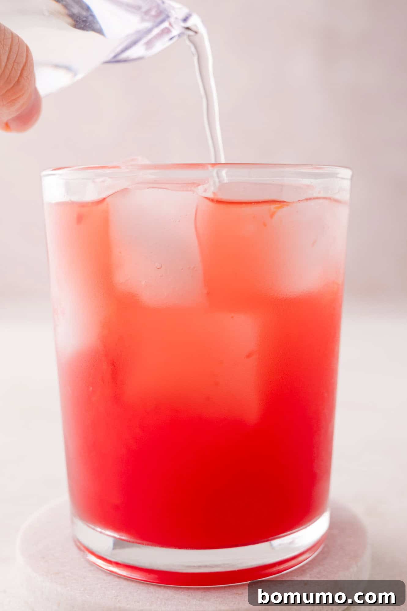 Crimson Sunset Sip 8 A hand carefully pouring clear sparkling liquid into a glass already containing ice and a red beverage.