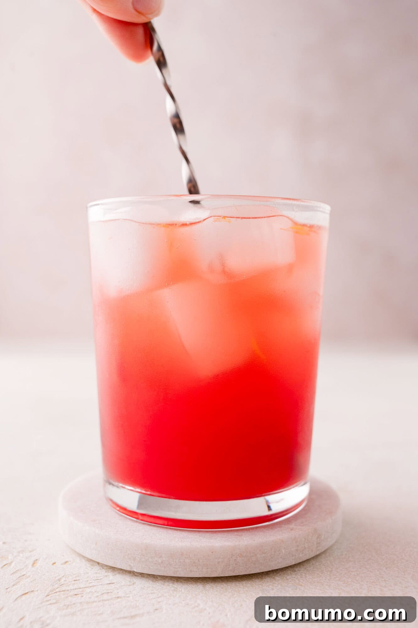 Crimson Sunset Sip 9 A hand gently stirs a red iced drink in a clear glass, resting on a round coaster, set against a clean, plain background.