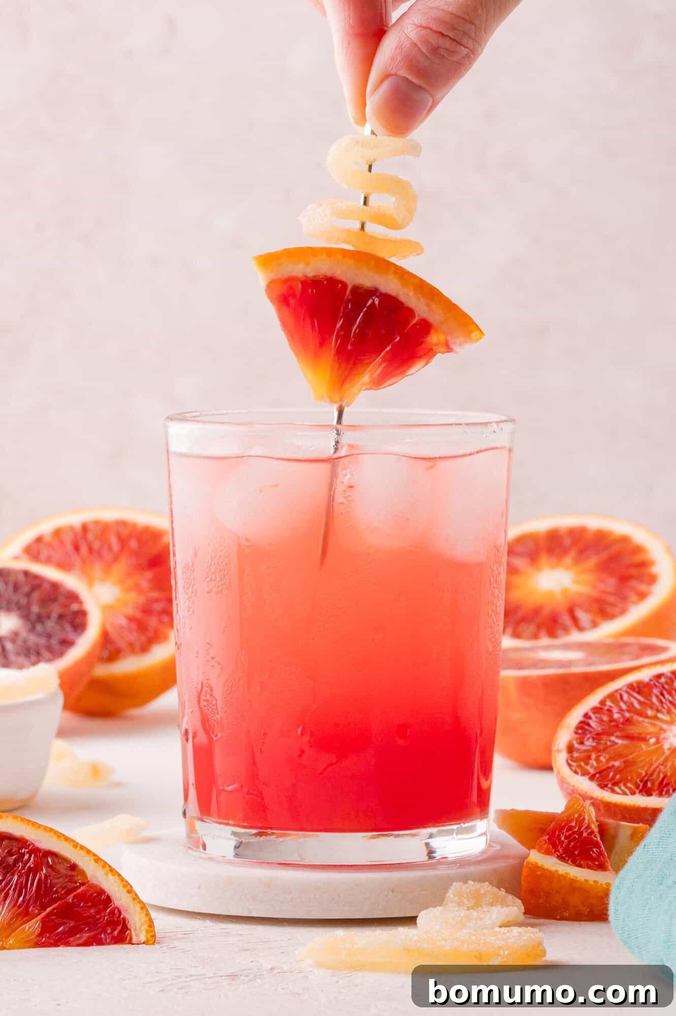 Crimson Sunset Sip 10 A hand elegantly placing a garnish of candied ginger and a blood orange slice over a glass of pink iced drink, surrounded by fresh blood orange slices.
